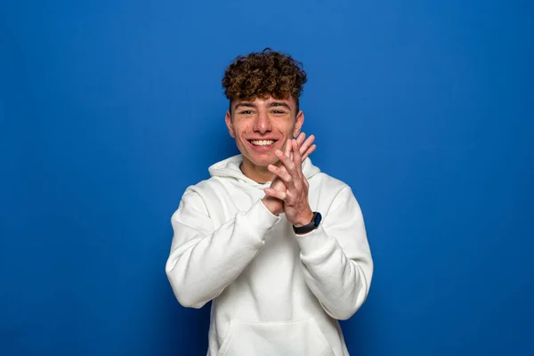 Caucasian young man with curly hair dressed in a white sweatshirt clapping hands smiling, isolated on blue studio background — ストック写真