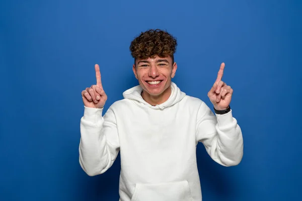 Portrait of a happy young man with curly hair pointing finger up at copy space isolated over blue background. — ストック写真