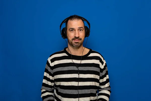 Hispanic man with a beard dressed in a striped sweater listening to music with headphones, isolated on blue studio background — Fotografia de Stock