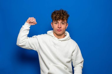 Kıvırcık saçlı, 20 'li yaşlarda beyaz kazak giyen, pazularını uzatan bir genç. Bu güç gösteriyor ki, Pastel mavi arka plan stüdyo fotoğraflarında izole edilmiş, insan yaşam tarzı konsepti.