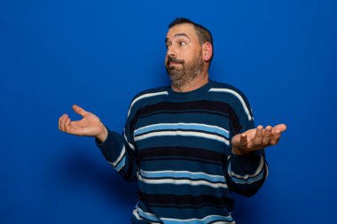 Çirkin çizgili süveter giyen, avuçlarını kaldırıp istifa jesti yapan, mavi arka planda soyutlanmış Latin sakallı bir adam.