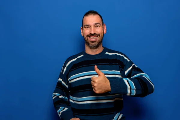 Bärtiger lateinischer Mann in gestreiftem Pullover, der zustimmend die Daumen nach oben zeigt, während er lächelt und in die Kamera blickt, isoliert über blauem Hintergrund. — Stockfoto