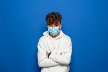 Caucasian young man with curly hair dressed in a white sweatshirt with his arms crossed and wearing a surgical mask to protect himself from the coronavirus, isolated on blue studio background