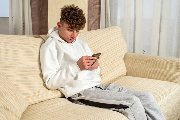 Caucasian young man with curly hair sitting on the sofa at home while consulting information on his smartphone with a very serious gesture. — Stock Photo, Image