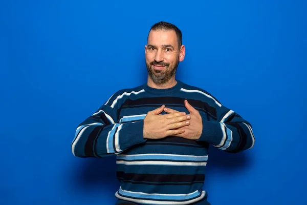 Handsome 40s man in a striped sweater put his hands folded on the heart isolated on blue background studio portrait. People emotions lifestyle concept. — Fotografia de Stock