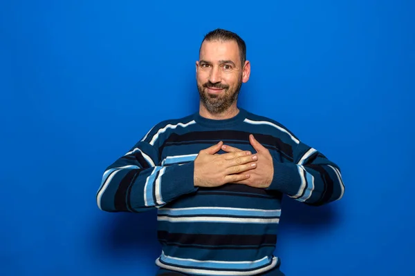 Bonito homem dos anos 40 com uma camisola listrada colocou as mãos dobradas no coração isolado no retrato do estúdio de fundo azul. Pessoas emoções conceito de estilo de vida. — Fotografia de Stock