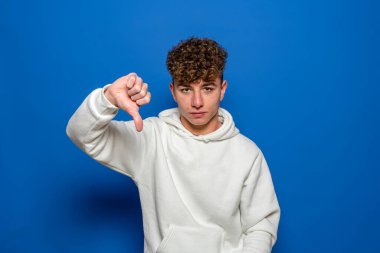 Mavi duvarlı arka plan stüdyo resminde izole edilmiş beyaz kazak giyen hoşnutsuz genç adam. İnsanlar gizli duygular ve yaşam tarzı konsepti geliştirdiler. Kopyalama alanı oluşturun. başparmağı göster.