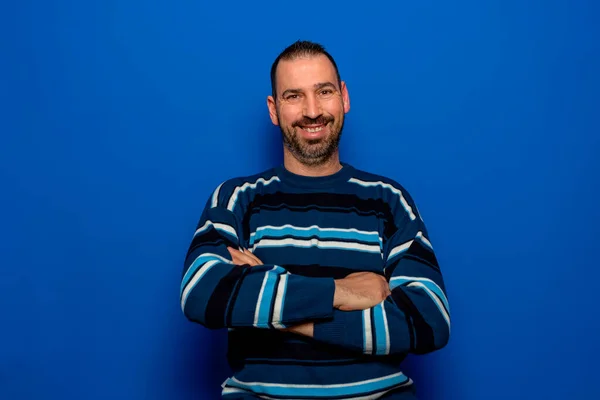 Portrait of bearded Latin man in his 40s with his arms crossed looking at the camera while smiling, isolated on blue studio background — Fotografia de Stock