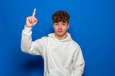 Beyaz kazaklı beyaz saçlı, beyaz saçlı beyaz bir genç işaret parmağıyla işaret ediyor. Mavi stüdyo arka planında izole edilmiş..