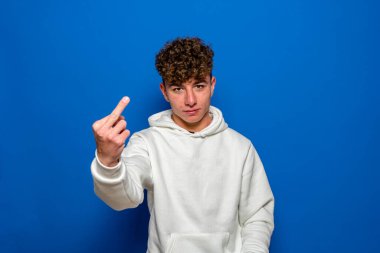 Young spanish man wearing white sweatshirt with curly hair standing over isolated blue background Showing middle finger, impolite and rude fuck expression