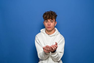Beyaz kazak giymiş, mavi arka planda izole edilmiş beyaz saçlı genç beyaz bir adam avuçlarıyla kameraya bir şey sunuyor..