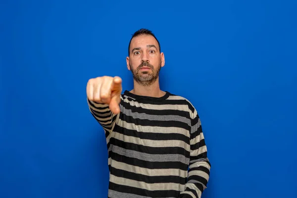 Portrait of handsome angry man pointing index finger at you on isolated blue background, looking at camera. Upset strict guy pointing index finger at camera choosing you.