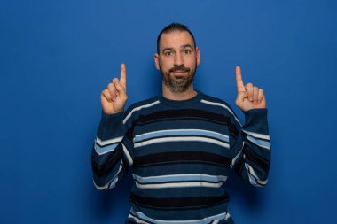 İspanyol sakallı adam çok çirkin çizgili kazak giyiyor. Mavi stüdyo arka planında, işaret parmaklarıyla gülümsüyor.