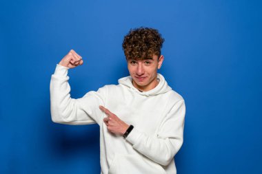 Kollarını gururla gösteren kıvırcık saçlı genç bir adam. Mavi stüdyo arka planında soyutlanmış.