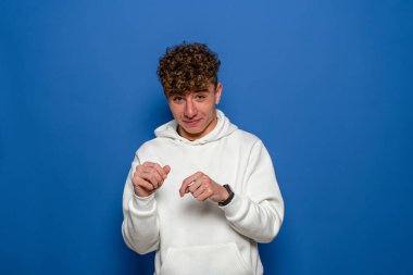 Nahoş bir genç adam portresi, garip bir durum, gergin bir şekilde ellerle oynuyor. Utanç verici. İzole edilmiş mavi arka plan. Olumsuz duygular yüz ifadesi hissi