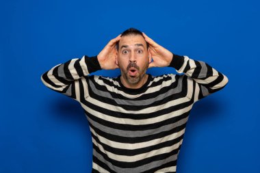 Stunned latin man with his head in his hands with a surprised expression isolated on blue background