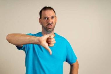 Young handsome man wearing casual blue t-shirt standing over isolated beige background looking unhappy and angry showing rejection and negative with thumbs down gesture. bad expression.