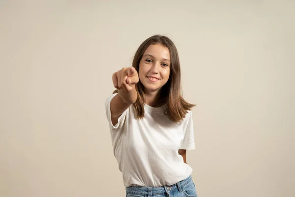 I choose you and command you. The smiling teenage girl pointing at camera, half-length close-up portrait on beige studio background. Human emotions, facial expression concept. Front view