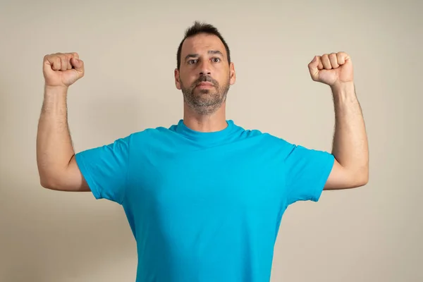 Caucasian happy unshaven strong sporty fitness student male 40s in casual blue t-shirt showing biceps muscles on hand isolated on beige background studio portrait.