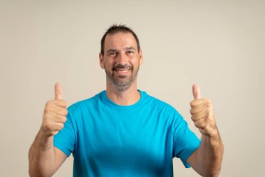 Bej arka planda izole edilmiş kameraya gülümseyen ve baş parmağını gösteren basit giyinmiş neşeli bir adamın portresi..