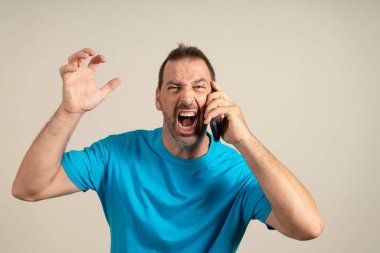 Pissed off bearded man yelling into his phone while on call with someone. He wears a blue t-shirt. Studio photo taken with beige background