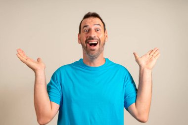 Mavi tişörtlü komik adam avuçlarını gösteriyor. Beklenmedik sürpriz jestler bej arka planda izole edilmiş. Çekici, mutlu adam iyi haberler duyuyor, bir şeyler yakalıyor..