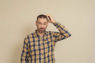 Ekose gömlekli, kafası kaşınan, bej bir arka planda izole edilmiş, çözüm arayan şüpheli sakallı İspanyol bir adam..