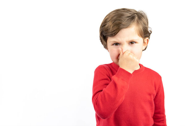 Beautiful boy in red sweater standing on white background isolated smelling something stinky and disgusting. Bad smell