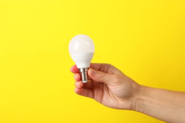 Kadın eli sarı arka planda enerji tasarrufu ampulünü tutuyor.