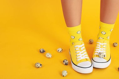 Çoraplı kadın bacakları, yumurtalı spor ayakkabıları ve sarı arka planda bıldırcın yumurtaları.