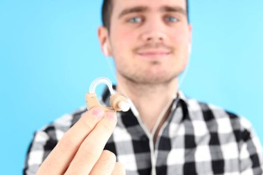 Mavi arka planda işitme cihazı tutan genç bir adam.