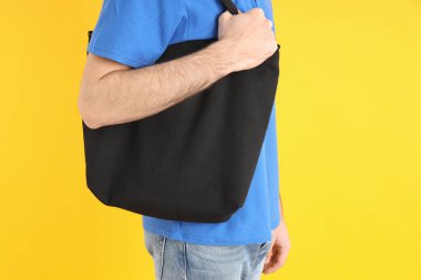 Sarı arka planda siyah çantalı mavi polo giyen adam.