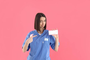 Female trainee nurse with pad and pregnancy test on pink background