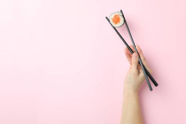 Kadın eli, pembe arka planda yemek çubuklarıyla suşi tutuyor.