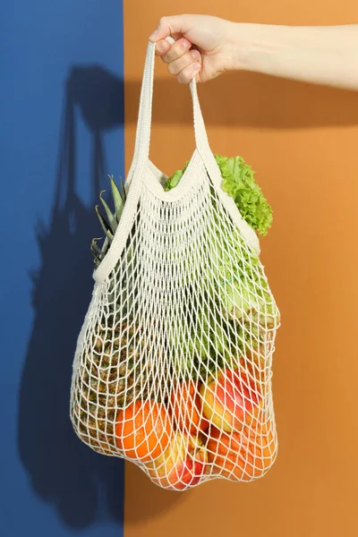 Female hand holds string bag with grocery on two tone background