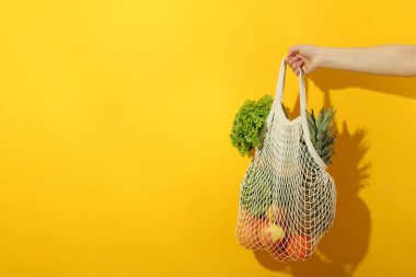 Kadın eli, sarı arka planda yiyecek dolu bir ip torbasını tutuyor.