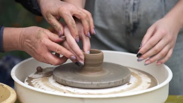 Una anciana artesana le enseña a una joven cómo hacer cerámica en una rueda de alfarero. Primer plano de las manos. — Vídeo de stock