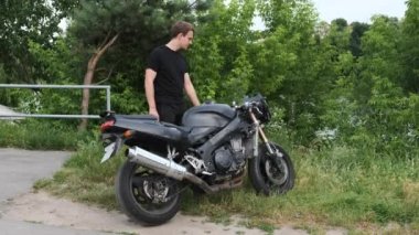 Siyah takım elbiseli genç bir adam ev yapımı siyah bir motosikletin yanında kollarını yeşil ağaçların arka planına doğru kavuşturuyor..
