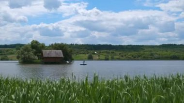 Gölün ortasında ahşap bir evi olan bir ada. Tahta bir salda bir adam kıyıya yüzmek için bir ip çeker. Güzel yaz manzarası.