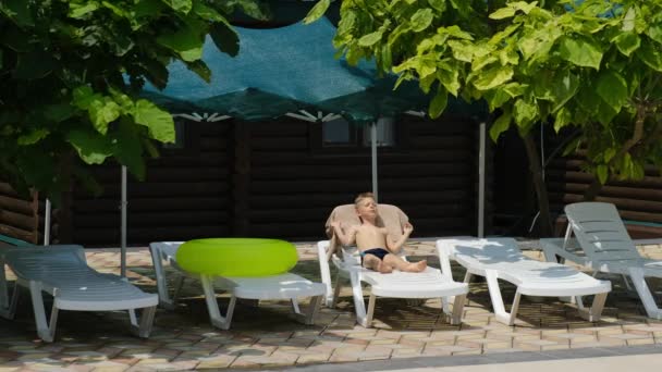 Menino Está Deitado Uma Chaise Longue Branca Tomando Sol Sol — Vídeo de Stock