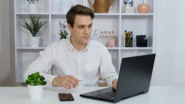 Genç bir adam ofisinde oturmuş kahve içiyor ve laptopuyla çalışıyor..