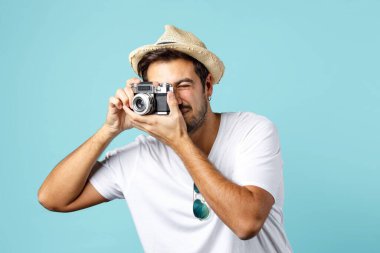 Beyaz tişörtlü ve yaz hasır şapkalı genç adam açık mavi arka planda izole edilmiş eski bir kamerayla fotoğraf çekiyor..