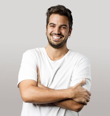 Beyaz tişörtlü, kollarını çapraz yapıp gri arka planda yaşayan yakışıklı bir adamın portresi..