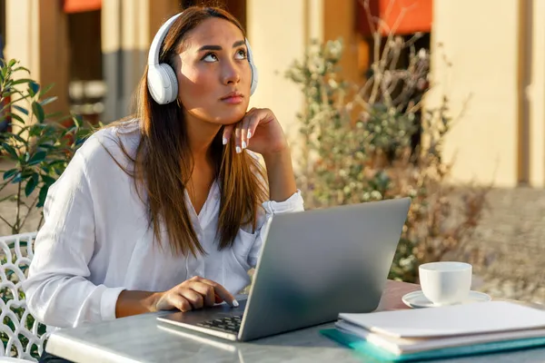 Pensive young woman wearing headphones and looking upwards while uses laptop. Pensive businesswoman or student leaning on chin meditates about future at a cafe table outdoor