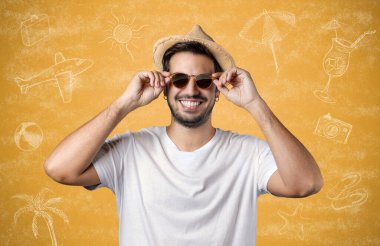 Genç sakallı adam yaz şapkası takıyor ve sarı bir arka planda izole edilmiş turistik tatil köyü çizimleriyle güneş gözlüğü deniyor. Tatil ve turizm kavramı.