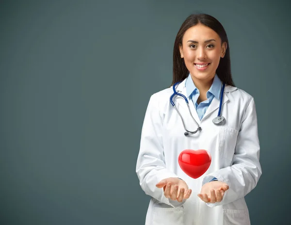 Portrait of smiling female doctor wearing white coat and stethoscope with a heart levitating above her hands and copy space. Concept of cardiology, organ donation and healthcare insurance. 3d illustration