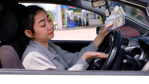 Desperate beautiful Asian woman holds cash dollar bills sitting inside her car at a gas station, concept of rising fuel prices
