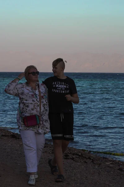 Мать-блондинка в легкой одежде гуляет по берегу моря со своим взрослым сыном. Летний вечер прогулка вдоль берега. Египет, Дахаб, 07.11.2021 — стоковое фото