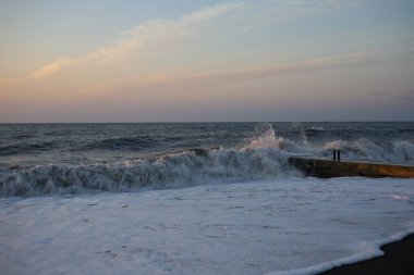 Şafak vakti kumlu bir sahilde fırtına. Hafif pembe gökyüzü ve köpüklü beyaz dalgalarıyla dramatik bir okyanus manzarası. Fırtınalı su ve sabah ışığında çarpan dalgalar.