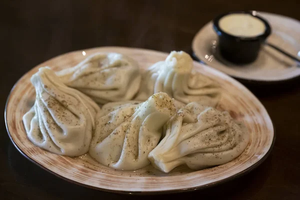 Khinkali - boulettes géorgiennes traditionnelles dans une assiette blanche avec sauce à la crème sure blanche sur une table en bois dans le restaurant — Photo
